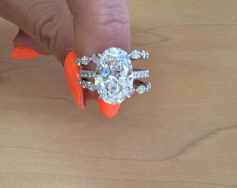Puede incluir: Un anillo de compromiso de diamante de forma ovalada con un engaste de tres bandas. La piedra central está rodeada de diamantes más pequeños, y las bandas también están engastadas con diamantes.