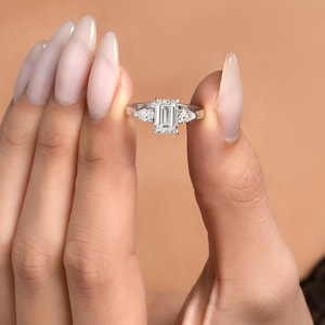 May include: A silver ring featuring a large emerald-cut diamond in the centre and two smaller pear-shaped diamonds on either side.