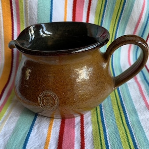 May include: A brown ceramic pitcher with a dark brown glaze on the inside and a gold logo on the side. The logo features a stylised rose and the text "Rose Creek Pottery".