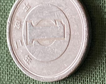 May include: A silver Japanese coin with visible characters and a central rectangular design. The coin is slightly off-center on a green textured surface.