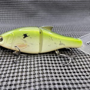 May include: A two-piece fishing lure in the shape of a fish. The lure is primarily light green with a white belly and a clear, translucent tail. It has a black spot and two silver hooks.