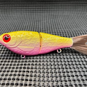 May include: A yellow and pink fishing lure shaped like a fish. The lure has a red eye and a brown tail. It has two metal hooks and is segmented in the middle.