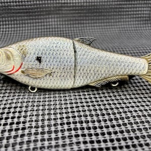 May include: A silver and gold-colored fishing lure shaped like a fish. The lure has red eyes, a red outline around the gills, and a segmented body. It features a detailed scale pattern and a hook for attaching to fishing line.