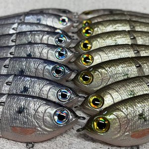 May include: A collection of silver and olive-green fishing lures shaped like fish. Each lure has a brown tail, detailed eyes, and a small hook. The lures are arranged in neat rows, suggesting they are ready for use in fishing.