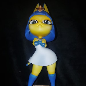 Op de afbeelding: Een geel kattenbeeldje met blauw haar en een witte jurk, staand op een bruine basis. De kat draagt een gouden ketting en heeft een blauwe hoofdtooi met een gouden ster.