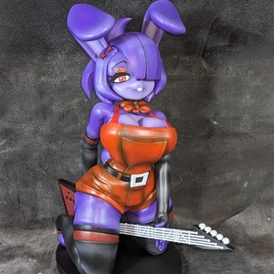 May include: Purple and orange anime-style figurine of a rabbit-like character holding a white electric guitar. The character is wearing a red bow tie and has red stars on its head.