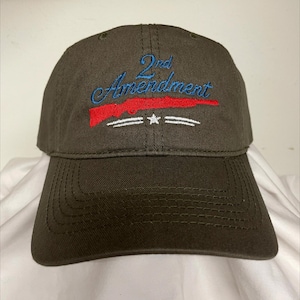 May include: A dark olive green baseball cap featuring embroidered text that reads 2nd Amendment in blue, a red rifle silhouette positioned below the text, and a white star flanked by two horizontal white lines at the bottom.