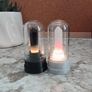May include: Two decorative glass cloches, each housing a glowing crystal. One has a black base and a dark crystal, the other a gray base and a white crystal. The crystals emit a warm, inviting light.