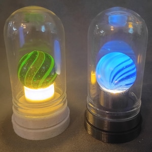 May include: Two decorative glass dome lights, each with a marble inside. One marble is green with a spiral design, the other is blue. The lights have a white and black base, respectively, and emit a soft glow.