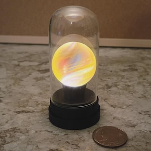 Puede incluir: Una cúpula de cristal decorativa con un orbe brillante multicolor en su interior. El orbe tiene remolinos amarillos, naranjas y azules. La cúpula se asienta sobre una base negra. Una moneda está al lado para comparar el tamaño.