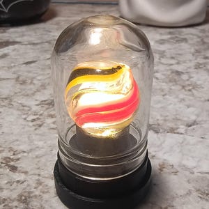 May include: A decorative glass marble with red, yellow, and white swirls, illuminated inside a clear glass cloche. The cloche sits on a black base, creating a unique lighting effect. The marble is approximately 1.5 inches in diameter.