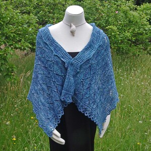 May include: A blue knitted shawl with a lacy pattern. The shawl is draped over a mannequin and is displayed against a green grassy background.