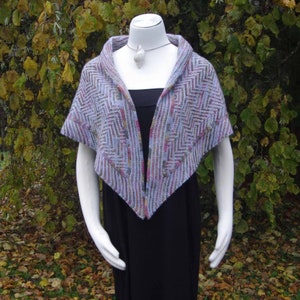 May include: A triangular shawl with a chevron pattern in shades of purple, pink, and gray. The shawl is draped over a white mannequin.