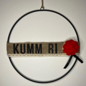 Puede incluir: Una corona de metal negro con una pancarta de arpillera que dice "KUMM RI" y una flor de fieltro rojo con una cinta negra.