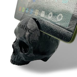 May include: A black skull-shaped phone stand with the word "CABLED" printed on the side. A tablet is resting on the stand.