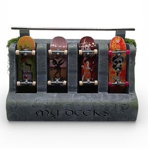 May include: A gray stone-like display stand holding four miniature skateboards. Each skateboard features unique graphics in various colors, including a skull, flames, and other designs. The stand has the text "my decks" engraved on the front.