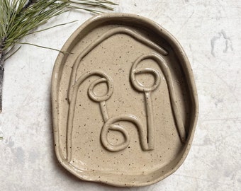 Puede incluir: Un plato de cerámica beige con un esmalte moteado. El plato tiene un diseño en relieve de una cara con dos ojos y una boca.