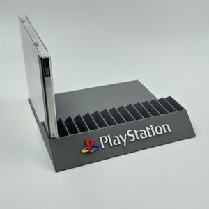 May include: A gray plastic PlayStation game case holder with slots for 15 games. The holder has the PlayStation logo and the word "PlayStation" printed on the front.
