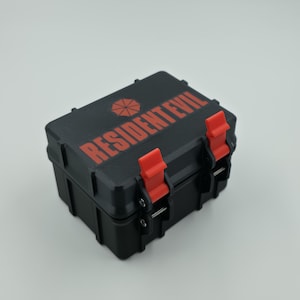 May include: A black rectangular case with red detailing and the words "RESIDENT EVIL" printed on the top. The case has two red latches and a red umbrella logo. The case is closed.