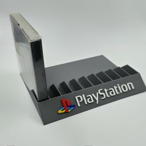 Puede incluir: Un soporte gris para juegos de PlayStation con múltiples ranuras, que sostiene una caja de juego. El logotipo de PlayStation es visible en la parte delantera. El soporte está diseñado para exhibir cajas de videojuegos.
