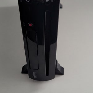 Puede incluir: Una consola Nintendo Wii U negra con una ranura para disco y un botón de sincronización rojo. La consola está de pie sobre dos patas negras.