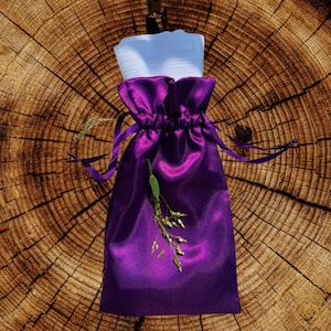 May include: A vibrant purple satin drawstring bag with a white item peeking out from the top. The bag is adorned with a sprig of green foliage. The background is a textured, brown wooden surface.