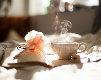 May include: A cozy scene featuring a steaming cup of coffee on an open book, with a delicate pink flower and reading glasses. A small heart-shaped cookie rests beside the cup. The soft light suggests a relaxing morning.