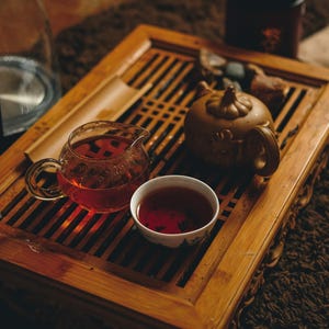 May include: A tea ceremony setup featuring a wooden tea tray with a glass pitcher, a small ceramic cup, and a teapot. The pitcher and cup are filled with a reddish-brown tea. A dark container sits in the background.