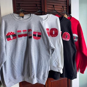 May include: Four sweatshirts in gray, white, black, and red, each with the word "OHIO" appliquéd in a patchwork design. The patchwork includes red, white, and black patterns. The sweatshirts are hanging on black hangers.