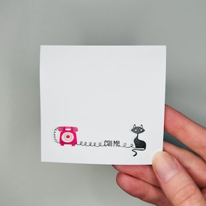 May include: A white square sticky note with a pink rotary phone illustration and a black cat illustration. The text "CALL ME" is between the phone and the cat. The note is held by a hand.