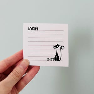 Könnte beinhalten: Ein weißer quadratischer Notizblock mit horizontalen Linien und den Wörtern "today's" und "to-do's" in Schwarz gedruckt. Eine schwarze Katzen-Silhouette befindet sich unten rechts. Der Notizblock wird von einer Hand vor einem hellblauen Hintergrund gehalten.