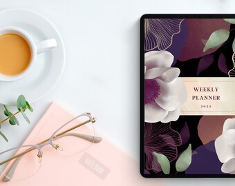May include: A digital tablet showing a floral weekly planner for 2023, with white and purple flowers on a black background. A cup of coffee, a pink notebook, and spectacles are arranged on a white surface, creating a workspace theme.