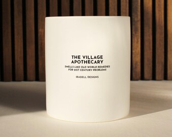 May include: A white cylindrical candle with the text "THE VILLAGE APOTHECARY" and "SMELLS LIKE OLD WORLD REMEDIES FOR 21ST CENTURY PROBLEMS" printed on the front. The candle is on a white surface with a wooden background.