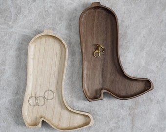May include: Two wooden trays shaped like cowboy boots. One tray is made of light-colored wood and holds three silver-toned rings. The other tray is made of dark-colored wood and holds a pair of small gold-toned hoop earrings. Both trays are hollowed out.