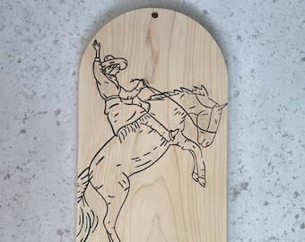 May include: A light-colored wooden plaque with an arched top and a small hole for hanging. The surface features a black line art illustration of a person in a cowboy hat riding a rearing horse. The text "cowtown. usa" is printed in the bottom right.