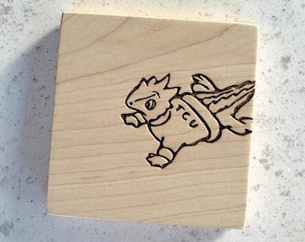 May include: A square light-colored wooden block featuring a dark laser-engraved illustration of a horned frog mascot wearing a jersey with the letters TCU printed on the front. The wood grain is visible across the surface of the square block.