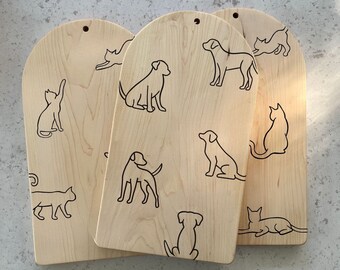 May include: Three light-colored wooden cutting boards with arched tops and small holes for hanging. Each board features black line art illustrations of various dogs and cats in different poses scattered across the smooth, grain-patterned surface.