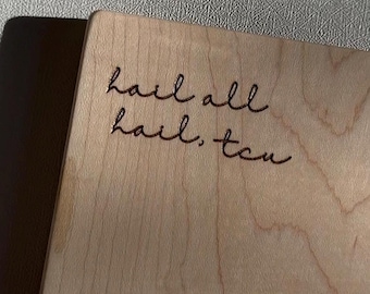 May include: A rectangular light-colored wooden board with a smooth grain pattern. The text "hail all hail, tcu" is engraved in a dark, cursive, handwritten-style font on the upper left portion of the wooden surface.