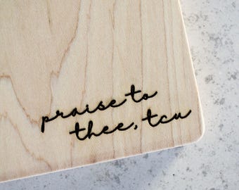 May include: A light-colored wooden cutting board with the phrase "praise to thee, tcu" in black script. The board has a smooth surface and a rounded corner. The background is a light gray, speckled surface.