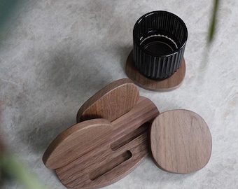 May include: A set of smooth, circular wooden coasters with a natural grain pattern, accompanied by a matching wooden holder featuring two slots for storage. A single black, vertically ribbed glass tumbler sits atop one of the wooden coasters.