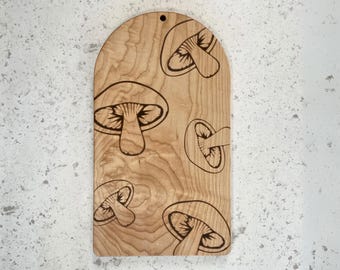 May include: A light brown wooden cutting board with a rounded top edge and a small circular hole for hanging. The surface features a laser-engraved pattern of five distinct mushrooms in varying sizes, showcasing the natural wood grain texture.