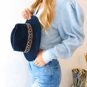 Può includere: Un cappello fedora nero con un bordo di perline dorate e blu. Il cappello è tenuto da una persona che indossa un maglione blu chiaro e jeans blu.
