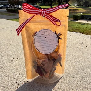 May include: A translucent brown pouch containing dried stovetop potpourri ingredients, tied with a red and white striped ribbon. A circular label reads: STOVETOP POTPOURRI SIMMER, Add all ingredients to a small pot, Add a few cups of water and simmer on low, Do not leave unattended and add water as needed, made with love.