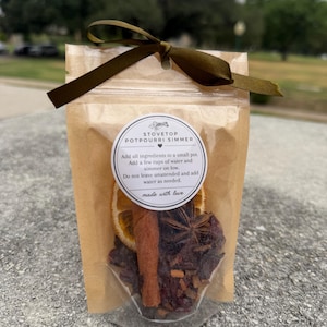 May include: A clear and brown pouch containing stovetop potpourri simmer ingredients, including a dried orange slice, a cinnamon stick, and star anise. A dark olive green ribbon is tied at the top. The label reads: STOVETOP POTPOURRI SIMMER.