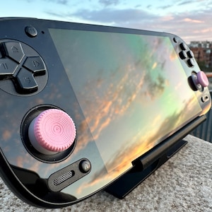 Pode incluir: Um console de jogos portátil preto com tampas de joystick rosa, repousando sobre um suporte preto. A tela reflete um pôr do sol colorido. O console possui um direcional e botões.