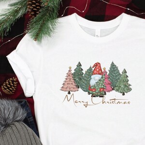 Könnte beinhalten: Weißes T-Shirt mit einem Weihnachtszwerg-Design. Die Grafik zeigt einen Zwerg mit roter Mütze und Bart, umgeben von Weihnachtsbäumen. Die Worte "Merry Christmas" sind in goldener Schrift unter den Bäumen geschrieben. Graue Turnschuhe und eine graue Mütze liegen neben dem Shirt.