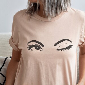 Può includere: Una maglietta rosa chiaro con una grafica di due occhi, uno dei quali sta strizzando l'occhio. Gli occhi sono neri e bianchi.