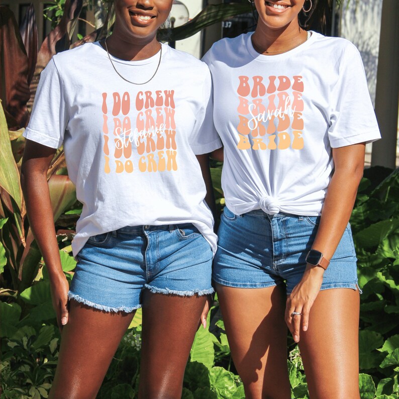 May include: Two women wearing white t-shirts with text printed on them. The woman on the left has a shirt that says "I Do Crew" in a retro font. The woman on the right has a shirt that says "Bride" in a retro font. Both women are wearing denim shorts.