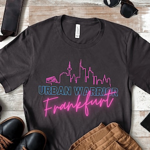 Puede incluir: Una camiseta gris oscuro con un diseño gráfico en rosa neón y azul. El gráfico presenta un horizonte de una ciudad con el texto "URBAN WARRIOR Frankfurt".