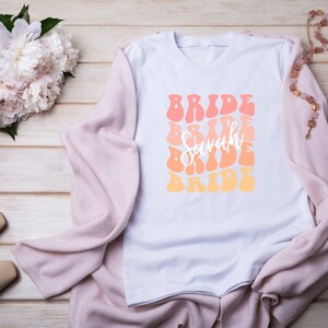 May include: White t-shirt with a retro-style graphic design that says "Bride" in a repeating pattern with the name "Sarah" in the center. The graphic is in shades of orange, pink, and yellow.